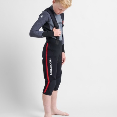 Criança vestindo calças de neoprene pretas com alças e detalhes em vermelho, com a palavra ROOSTER na lateral.