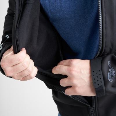 Casaco preto com fecho e logotipo SOFTSHELL no punho, vestido sobre pessoa com t-shirt azul