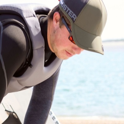 Homem com boné com texto 'MONSTER' e colete salva-vidas cinzento junto à água