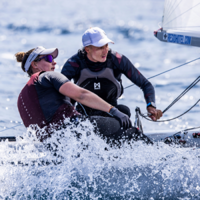 Dois velejadores em barco pequeno com coletes e bonés num mar agitado