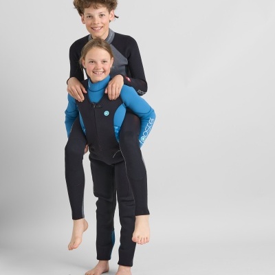 Duas crianças com fatos de neoprene para desportos aquáticos, uma a carregar a outra às costas, fundo cinzento.