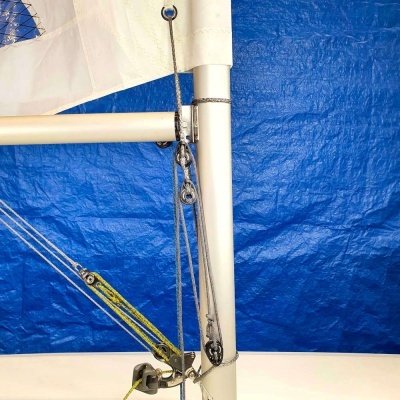 Equipamento de vela com mastro branco, cordas, polias e pano branco, fundo azul.