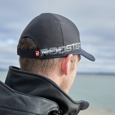 Homem visto de costas usando boné preto perfurado com 'ROOSTER' e casaco escuro junto à água