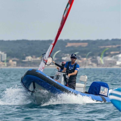Barco insuflável azul com homem a bordo em mar calmo e velas ao fundo