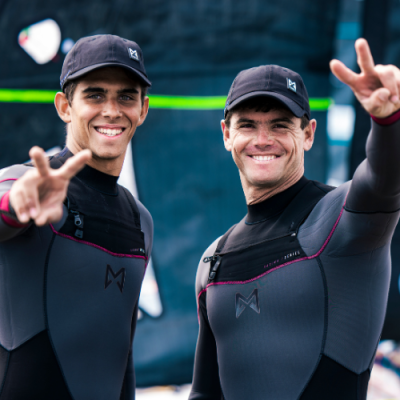 Duas pessoas com fatos de neoprene pretos e bonés pretos fazem sinal de paz