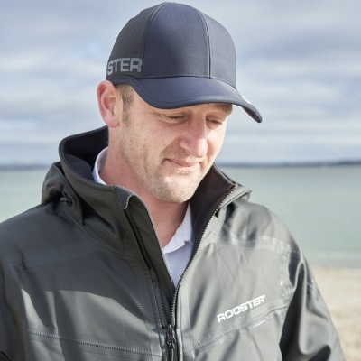 Homem com casaco cinza e chapéu de basebol cinza azulada com logótipo ROOSTER na praia