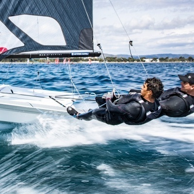 Dois velejadores em fato de neoprene preto a velejar numa pequena vela branca com vela preta na água azul.