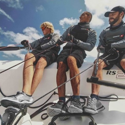 Três homens com roupa desportiva a velejar num barco RS sob céu azul nublado.