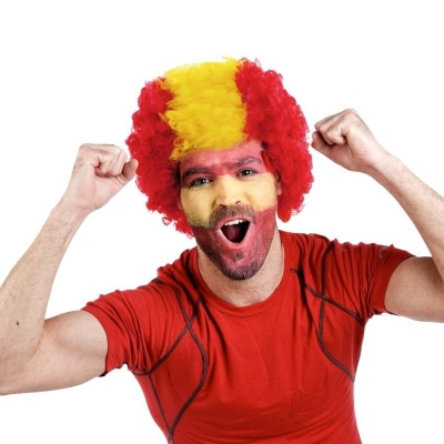 Homem com peruca cacheada vermelha e amarela, rosto pintado e t-shirt vermelha, comemora com os braços erguidos