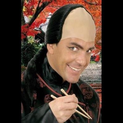 Homem com máscara de testa grande e cabelo preto sorrindo e segurando pauzinhos para comida, com fundo de árvores vermelhas e escadaria