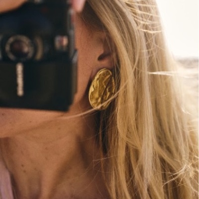 Brinco dourado grande e texturizado em mulher loira com câmera fotográfica preta