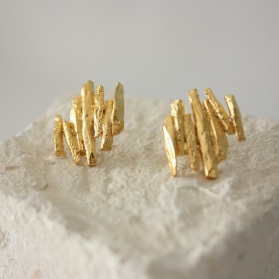 Brincos dourados com textura irregular sobre bloco branco rugoso.