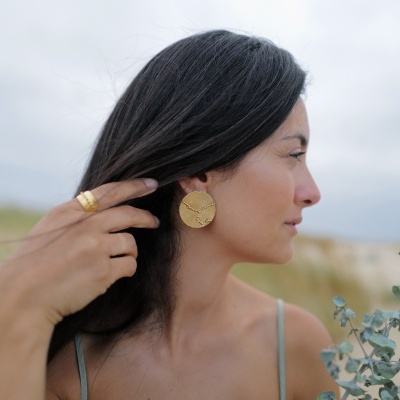 Mulher de perfil com brincos e anel dourados segurando planta seca em praia