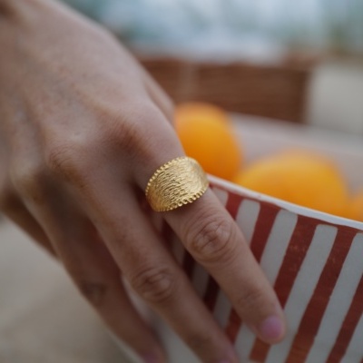 Anel dourado texturizado num dedo próxima a uma tigela listrada com laranjas.