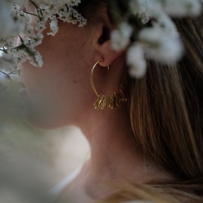 Brincos dourados de argola com pendentes numa orelha feminina rodeada de flores brancas
