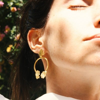 Brincos dourados texturizados em forma oval num rosto feminino com olhos fechados ao ar livre