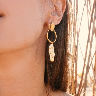 Brinco dourado pendente usado por mulher com cabelo castanho claro e fundo com ramos secos