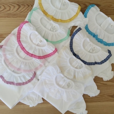 Bodies brancos de bebé com golas coloridas e rendadas várias cores sobre fundo de madeira.