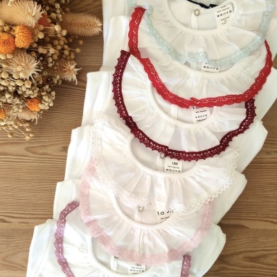 Sete bodies brancos para bebé com golas em renda colorida empilhados sobre madeira ao lado de flores secas