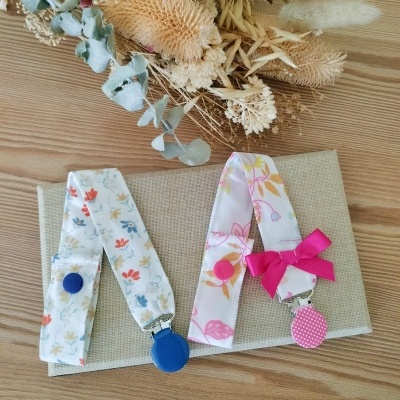 Dois prendainfantis de tecido floral com clips coloridos sobre tecido bege e fundo de madeira clara com flores secas