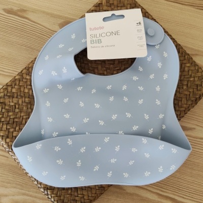 Babete de silicone azul claro com padrão de folhas brancas e etiqueta da marca tutete