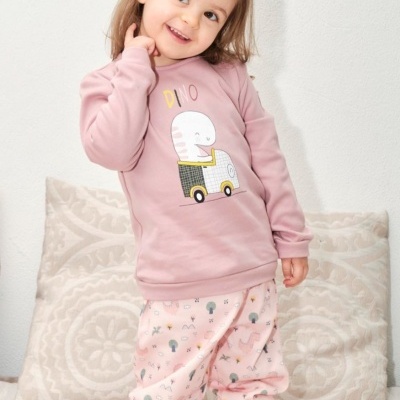 Menina com pijama rosa com estampa de dinossauro e calças estampadas, posando em sofá com almofadas.