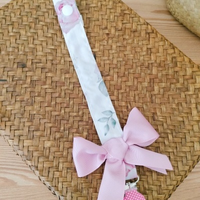 Prendedor de chupeta branco floral com laço rosa e clip rosa com bolinhas sobre palha trançada