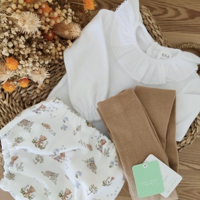 Conjunto de roupa para bebé com camisa branca, calças curtas com padrão e meias castanhas sobre superfície de madeira com flores secas