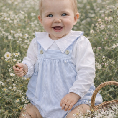 Bebé sorridente com vestido azul e camisa branca sentado num campo de flores com cesto de vime.