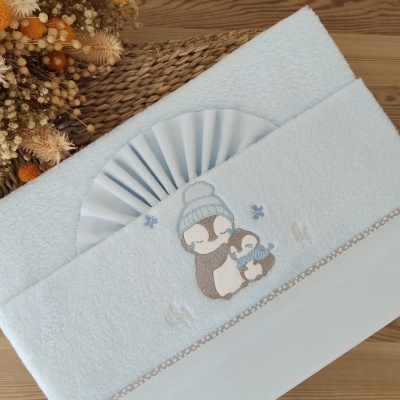 Toalha de bebé azul clara com bordado de pinguins e flores secas ao lado