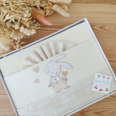 Conjunto de roupa de bebé bege com coelho bordado, embalagem branca e flores secas ao lado