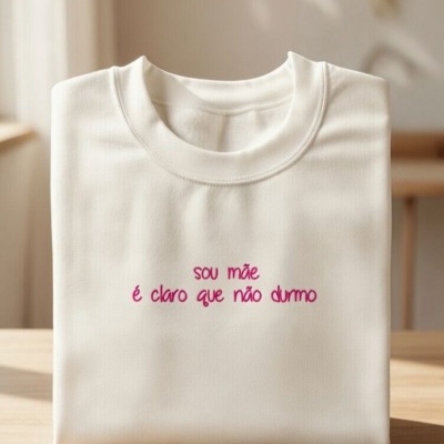 T-shirt branca dobrada com texto bordado rosa sobre mesa de madeira clara