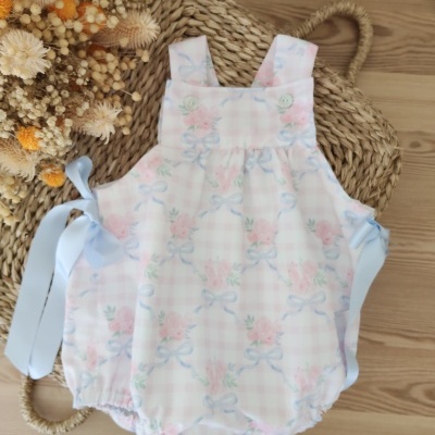 Body de bebé rosa claro com flores e laços azuis, botões frontais e fitas azuis, sobre esteira de palha