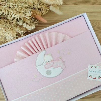 Jogo de cama rosa para bebé com bordado de urso e lua numa caixa branca sobre mesa de madeira