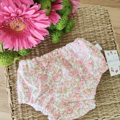 Cueca de bebé floral em tons rosa e verde sobre superfície de rattan com flores cor-de-rosa ao lado