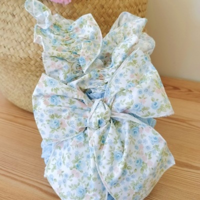 Vestido infantil floral com laço grande, sobre mesa de madeira clara e cesto de palha ao fundo