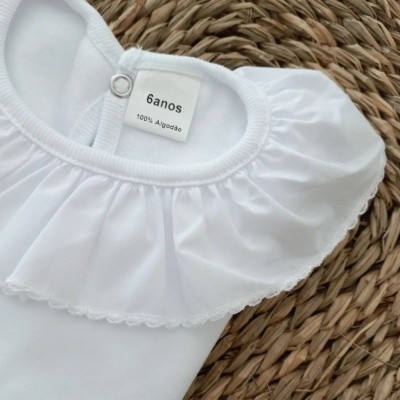Body branco para bebé com gola franzida e etiqueta 6 anos sobre cesta de palha