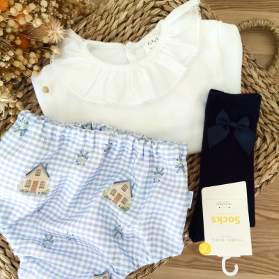 Conjunto de roupa de bebé com camisola branca, calções azul claro com padrão de casas e flores, meias pretas com laço.