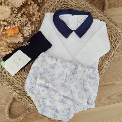 Conjunto de roupa de bebé com camisa branca, colarinho azul, cueiro com padrão de coelhos e flores azuis e meias pretas em cesta de verga