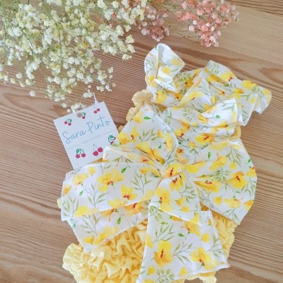Roupa de bebé amarela com laços e flores, cartão Sara Pinto e flores secas sobre madeira