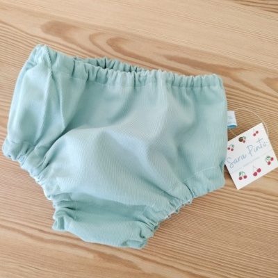 Calção de bebê verde água com elásticos, etiqueta Sara Pinti, sobre mesa de madeira clara