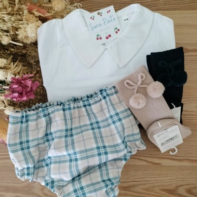 Conjunto de roupa infantil com camisa branca, calção aos quadrados azul e branco, e duas meias coloridas em superfície de madeira com flores secas.