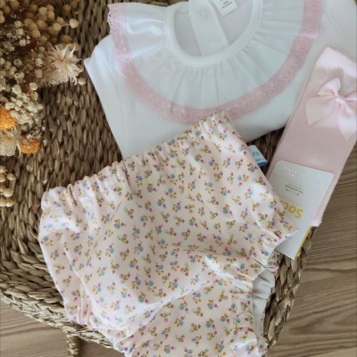 Conjunto de roupa para bebé com body branco e gola rosa, calções florais e meia rosa com laço sobre cesto de fibra natural.