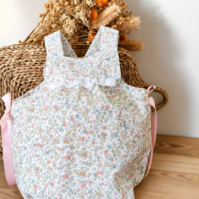 Macacão infantil floral com renda branca e fitas cor-de-rosa sobre cadeira de vime com flores secas.