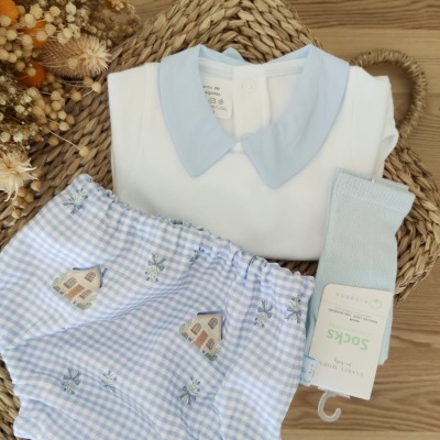 Conjunto de roupa de bebé branco e azul claro com padrão de casas e meias azuis sobre base rattan