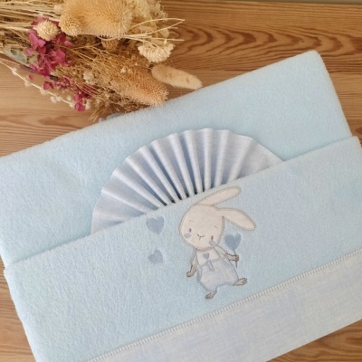 Toalha azul clara com bordado de coelho e leque branco numa superfície de madeira com flores secas ao lado