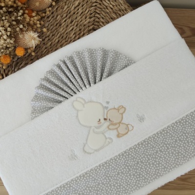 Toalha branca com bordado de coelhos e acabamento cinzento de bolinhas em superfície de madeira com flores secas e vime