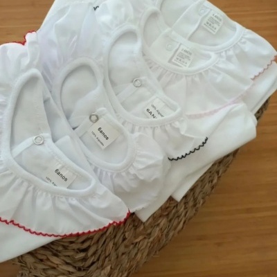 Bodies de bebé brancos com gola bufante e bordas coloridas numa cesta sobre madeira