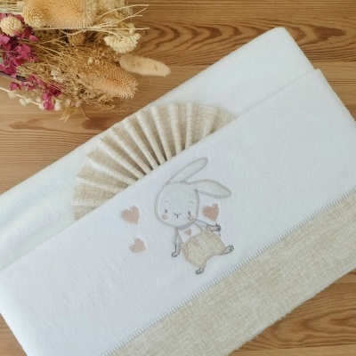 Toalha para bebé branca e bege com bordado de coelhinho e corações, decorada com laço bege, ao lado de flores secas