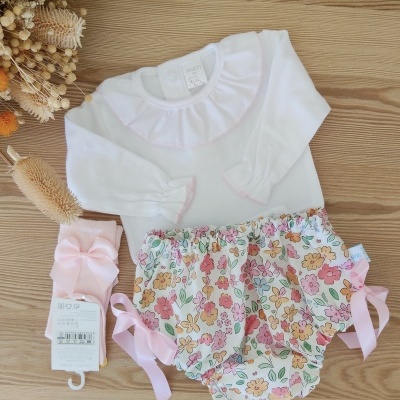 Conjunto de bebé com body branco, calções florais e meias rosas com laço sobre mesa de madeira.